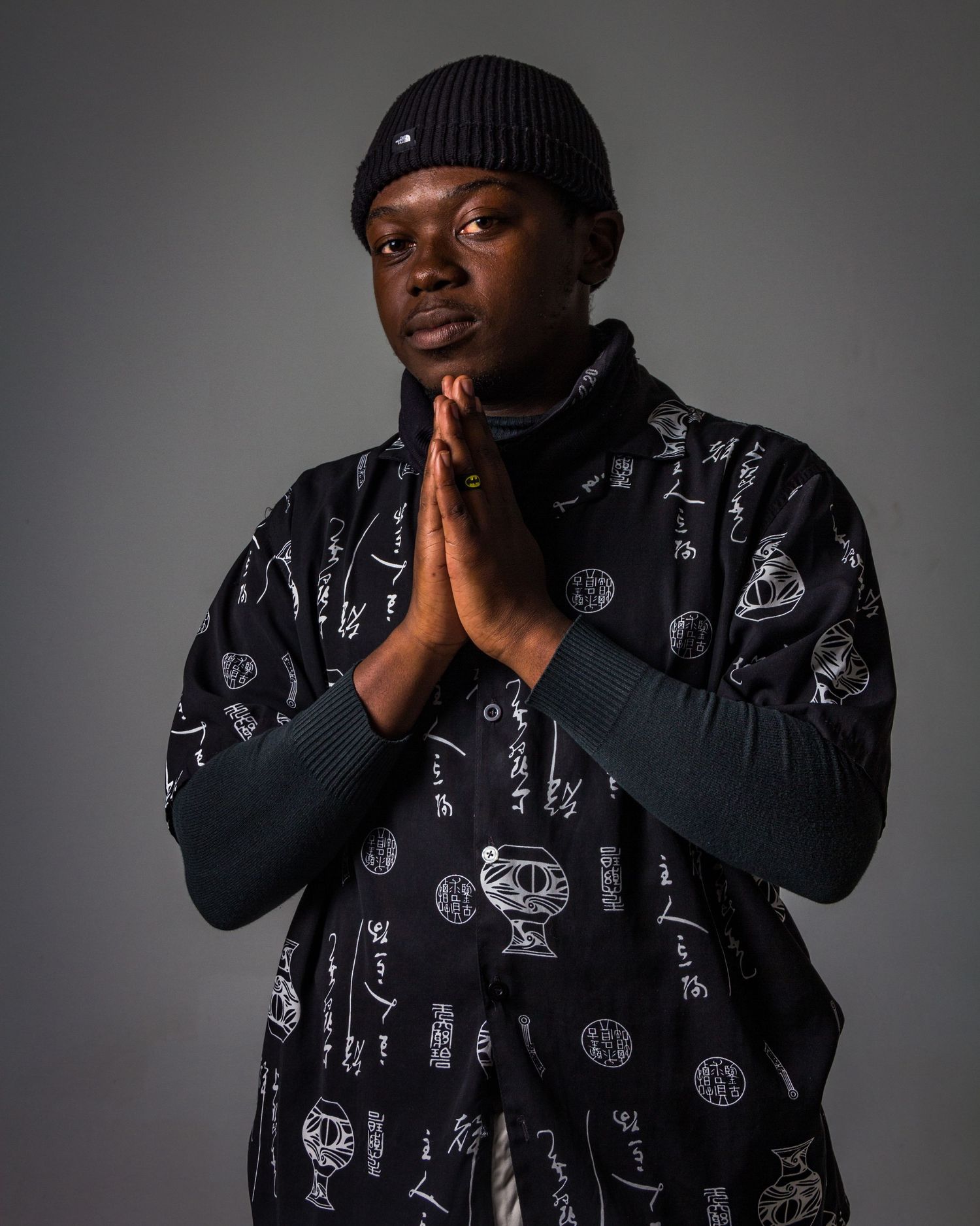 Person wearing black patterned jacket poses thoughtfully against gray background in dramatic studio lighting.