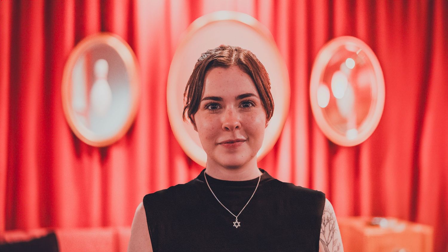 Three people stand in front of a bright red curtain backdrop with clocks visible on the wall behind them.