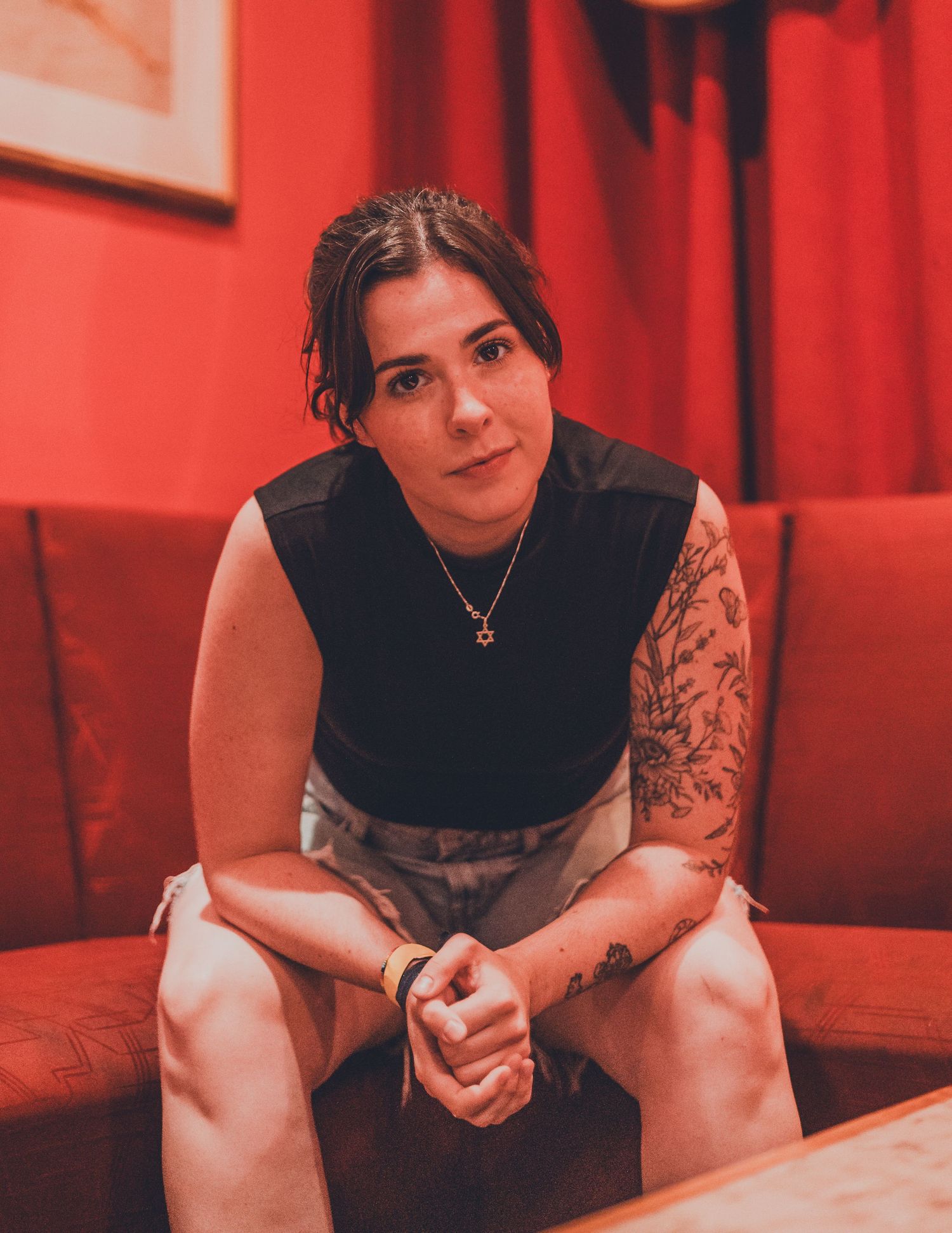 Person in black sleeveless top sitting on floor in red-tinted room with tattoos on arm.