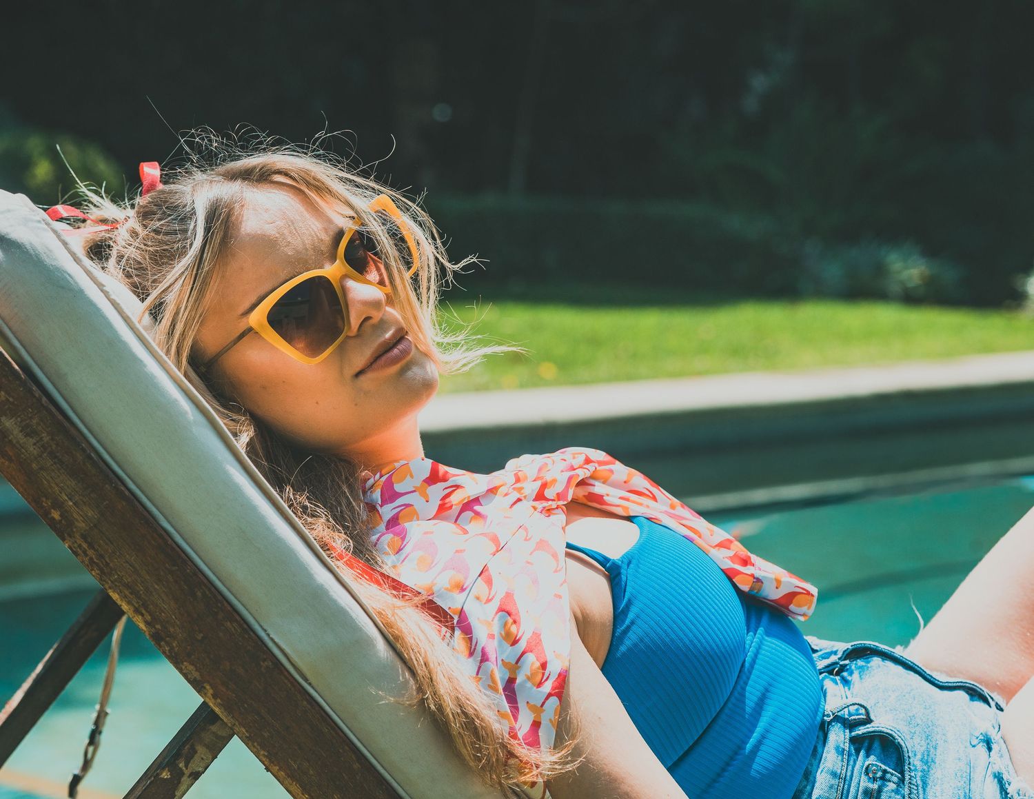 Person in sunglasses and colorful shirt relaxing on a lounge chair by a pool on a sunny day.