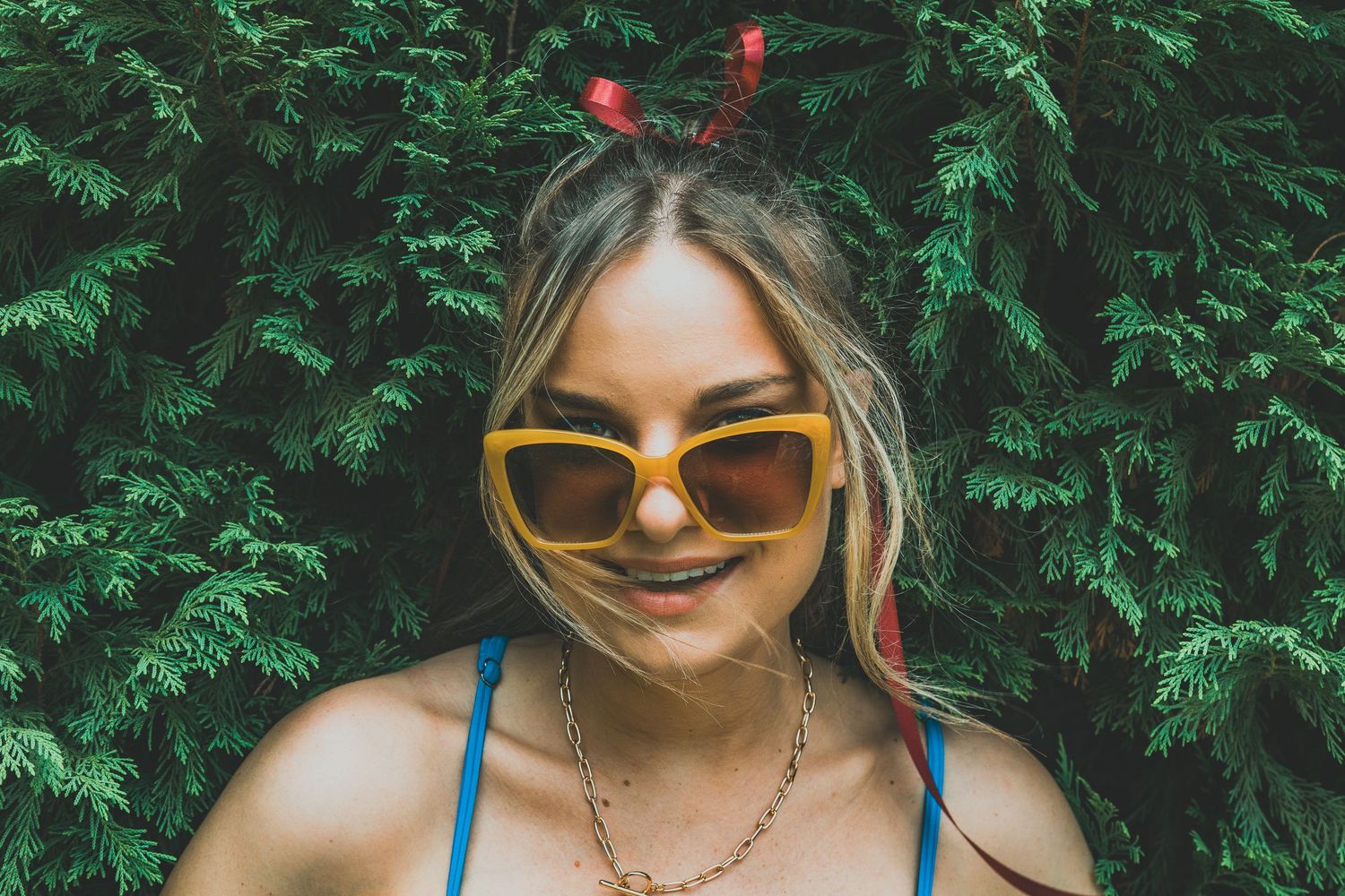 A person in sunglasses and a blue tank top poses against lush green foliage on a sunny day.