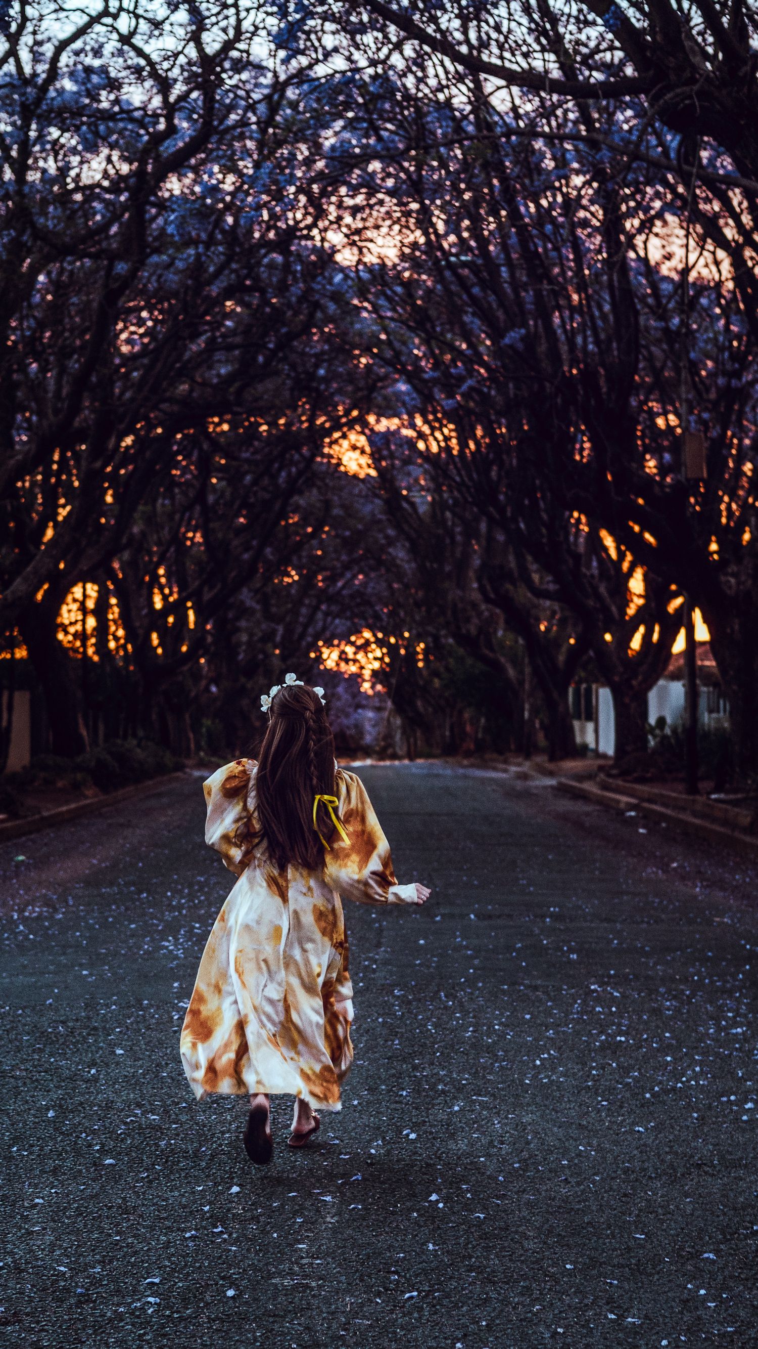 A figure in a flowing dress walks down a tree-lined road at sunset creating a dreamy silhouette against the orange sky.