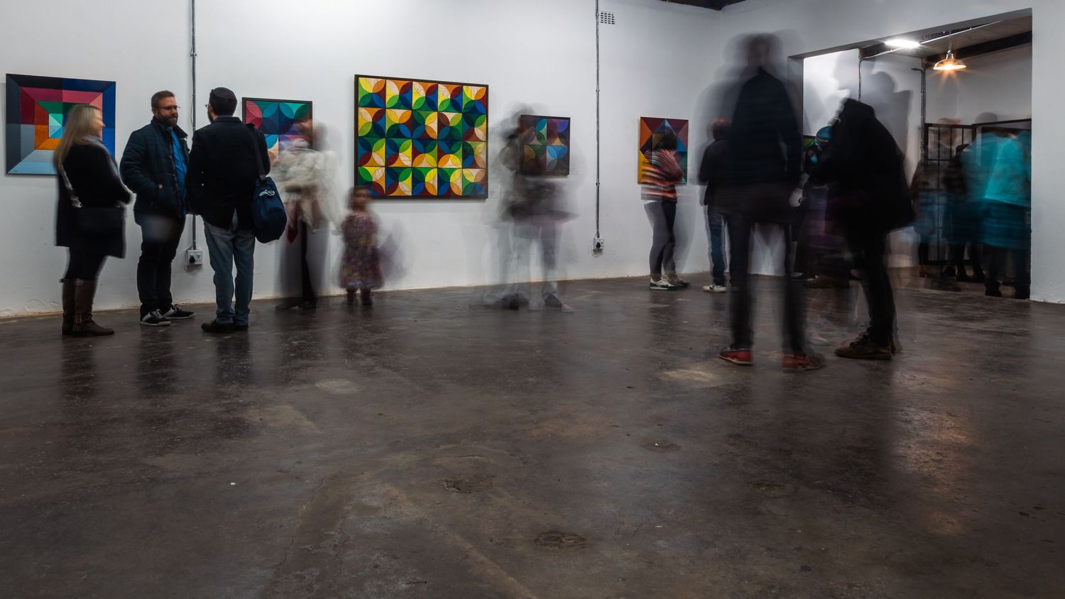 Panoramic view of an art gallery exhibition space showing colorful geometric paintings on white walls.