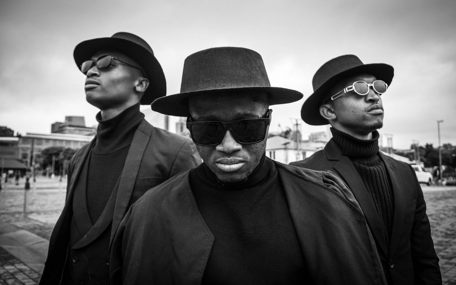 Three men in black turtlenecks and hats pose together in a dramatic black and white portrait.