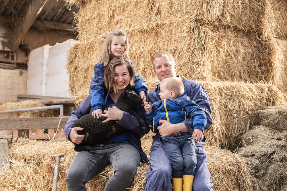 Family sits together laughing on hay bales at a farm with two young children in casual attire.