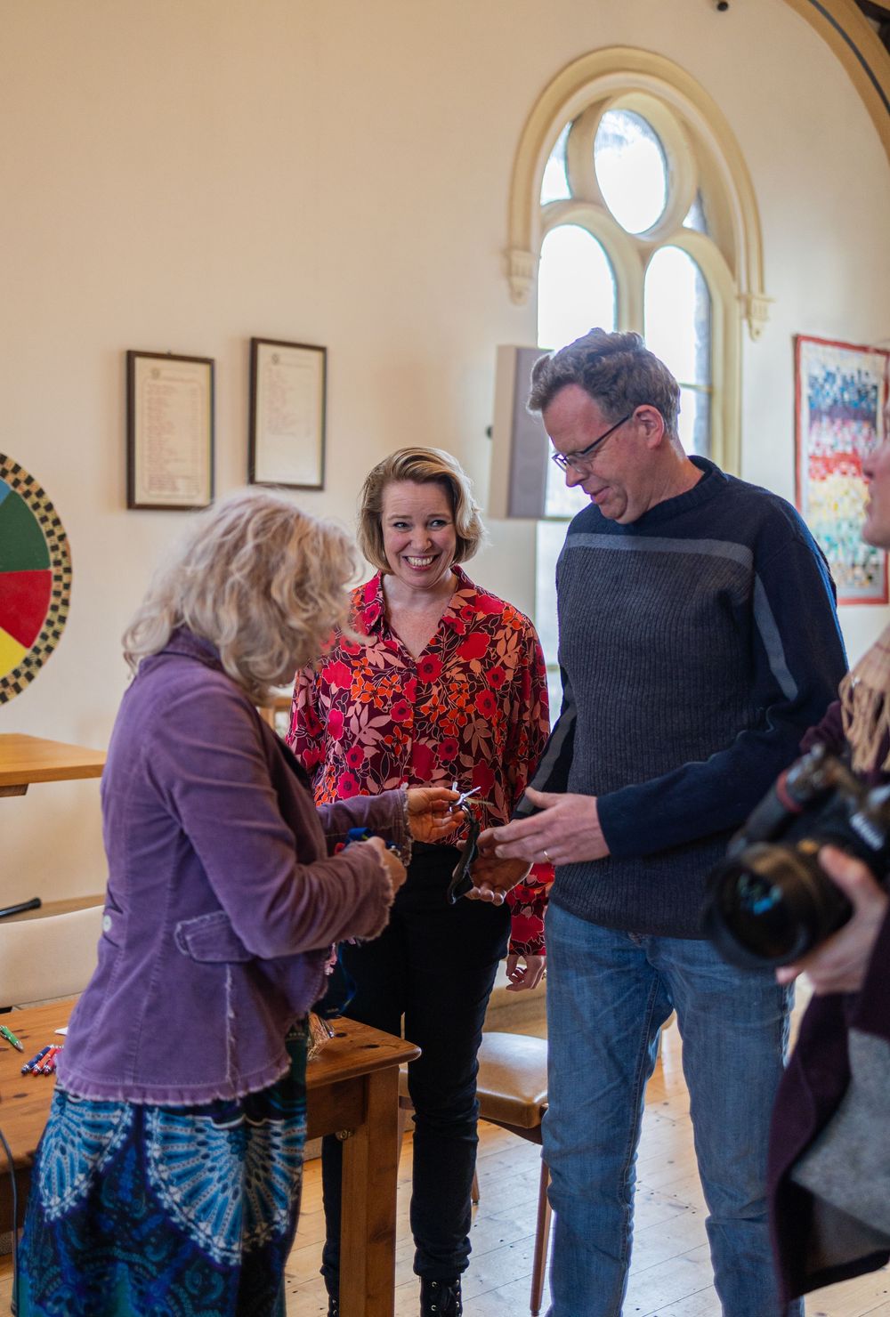 Three people examine something together in a room with gothic arched windows and framed artwork.