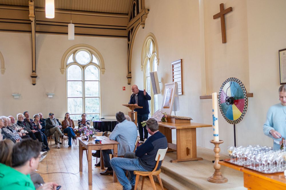 People gathered in a church hall with gothic windows and a wooden cross on the wall.