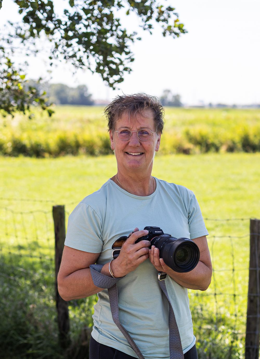 A photographer in a white t-shirt smiles while holding a camera in a sunny field with trees in the background.