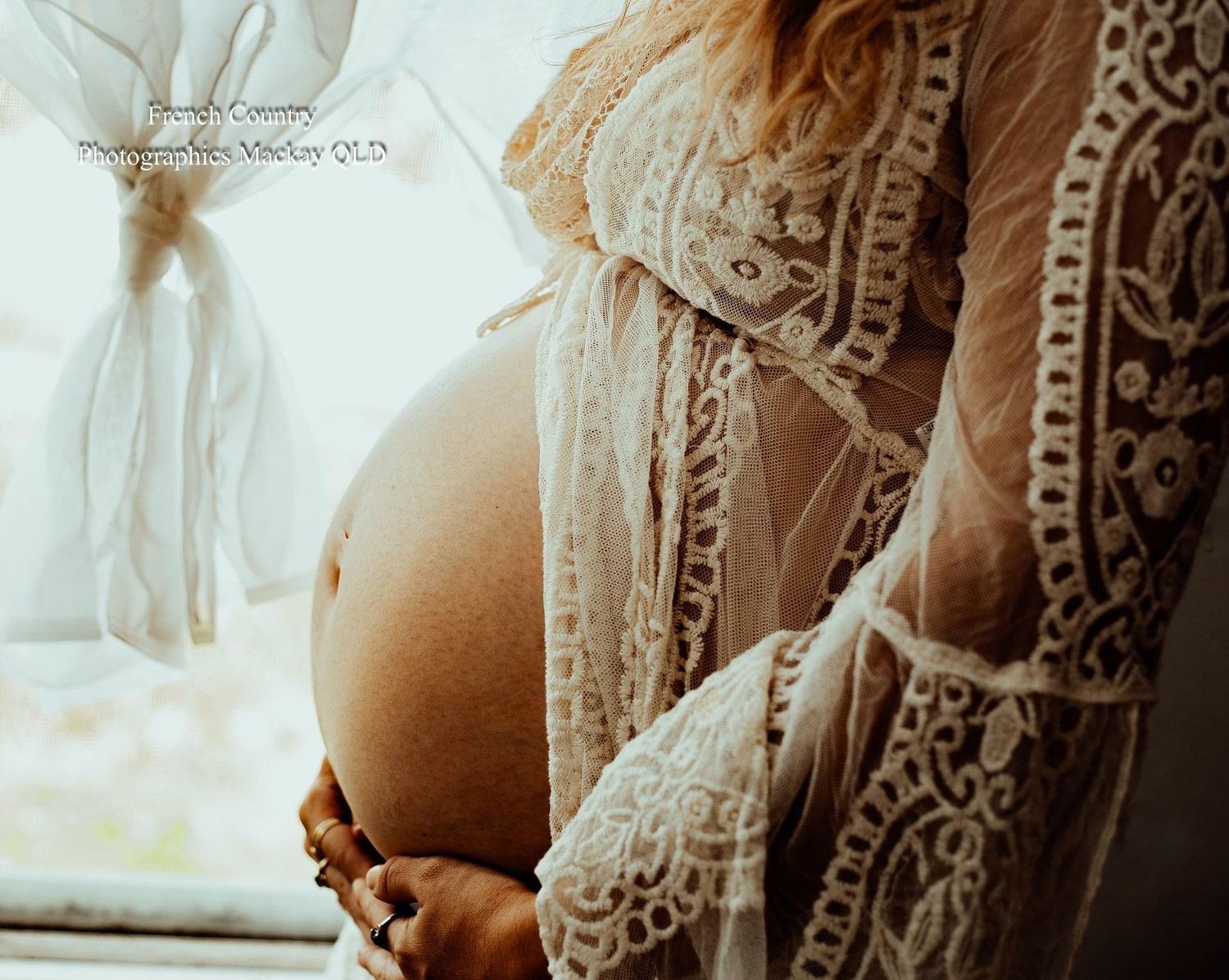 Close-up of pregnant belly draped in delicate white lace against window light.