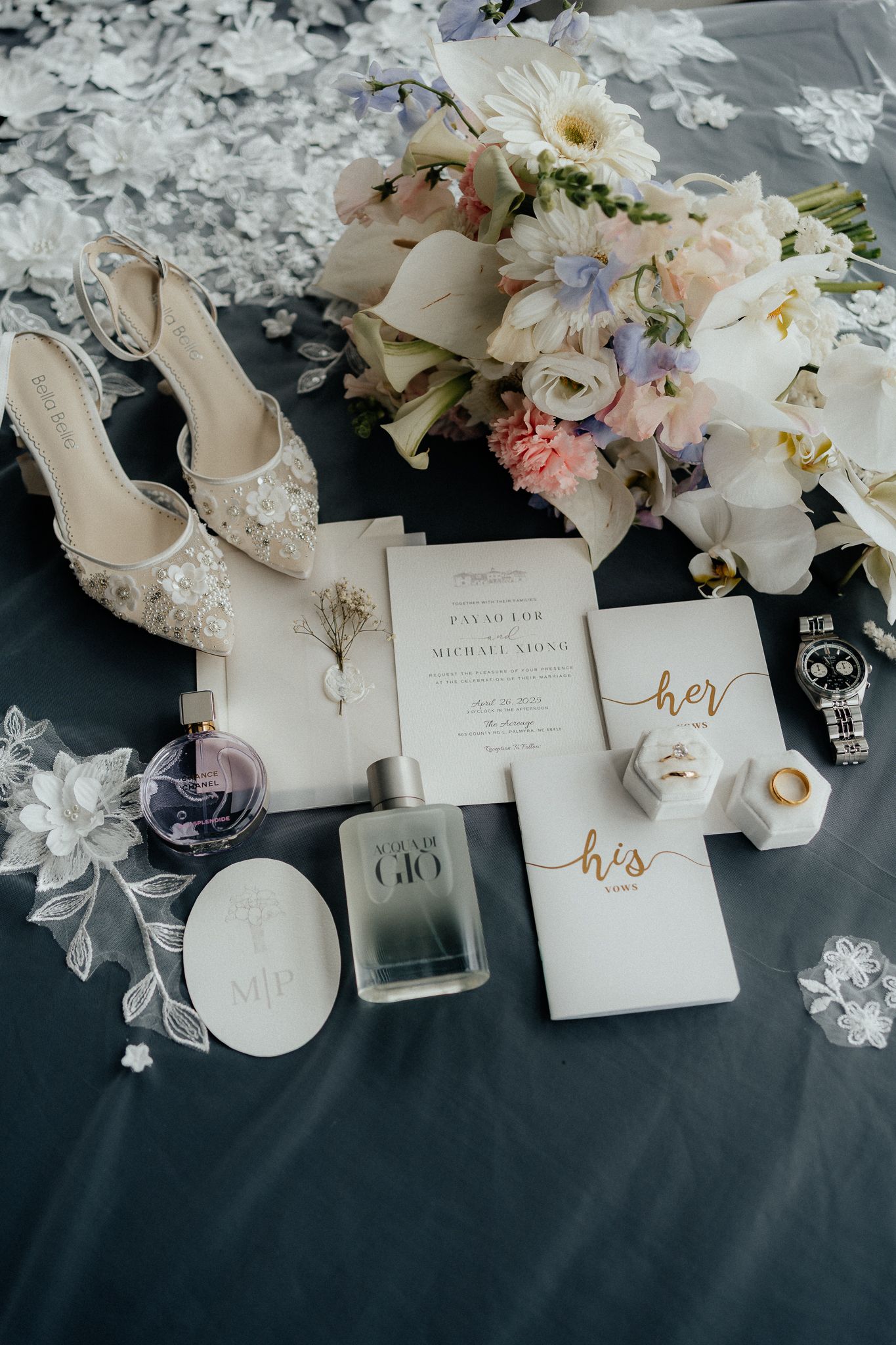 Wedding details flatlay with bridal shoes, invitations, perfume and floral arrangements on dark fabric.