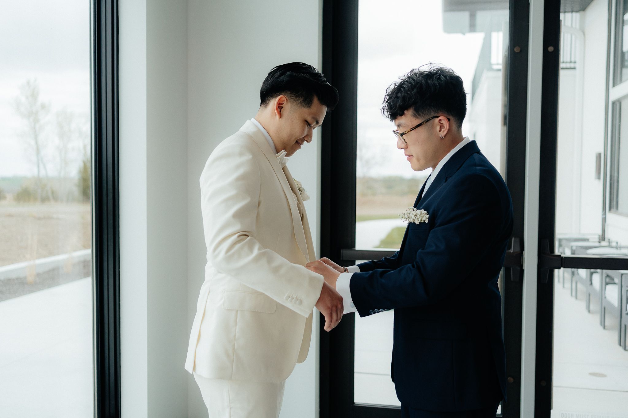 A groom in white suit gets help with final adjustments from his groomsman in a black suit by a window.