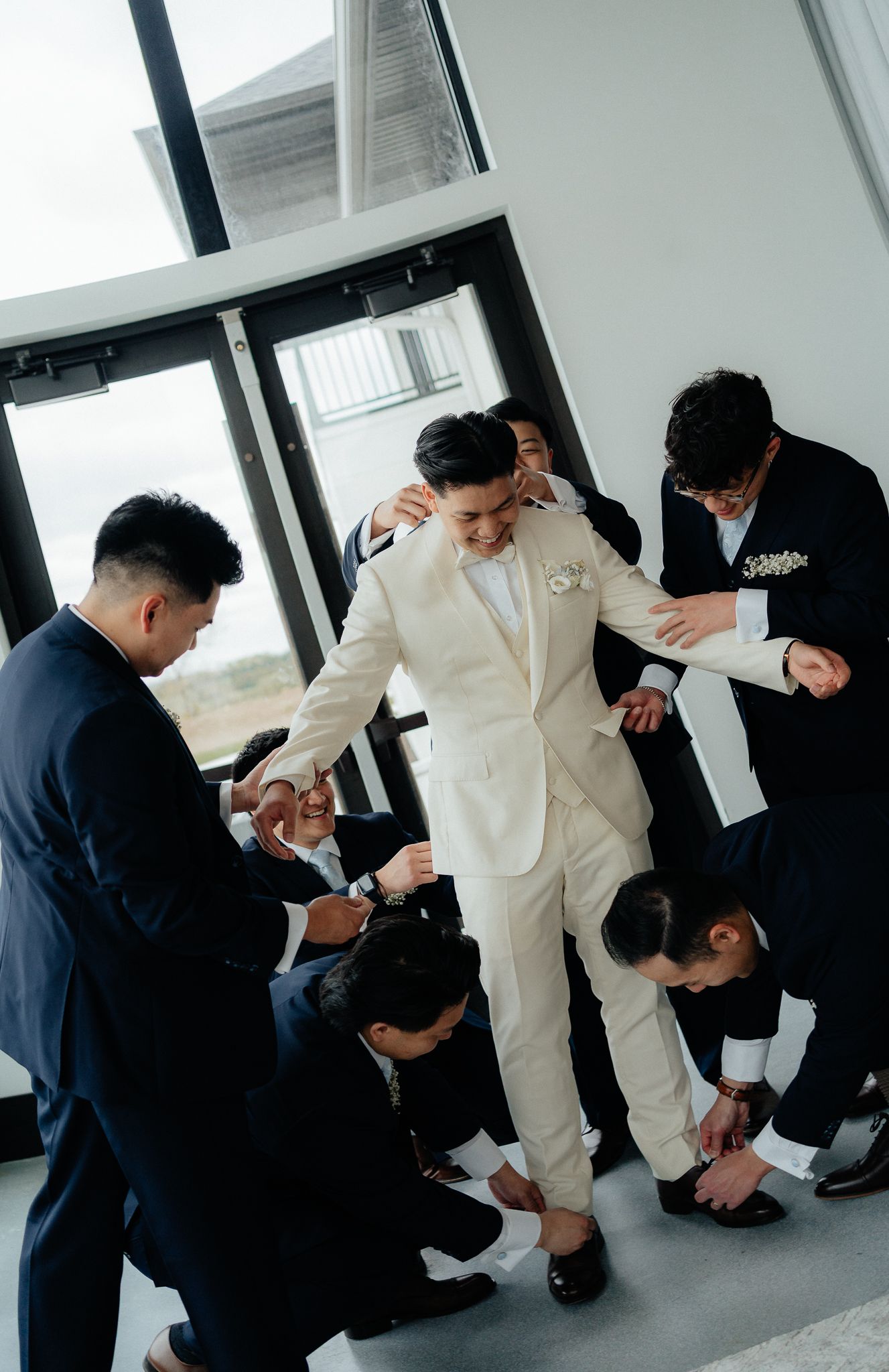 Groomsmen help adjust the shoes and attire of a groom wearing a white suit in a brightly lit room.