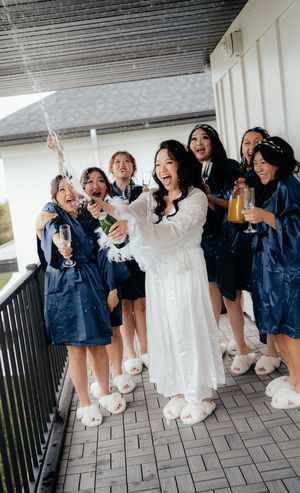 Bridesmaids in navy robes celebrate with champagne alongside bride in white on a balcony.