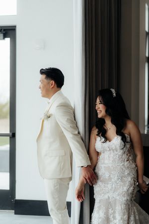A wedding couple in white attire stand back to back in a bright modern venue.