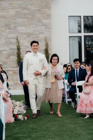 A groom in cream suit walks down the aisle during an outdoor ceremony.