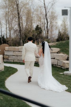 Newlyweds walk along a stone-lined path with a flowing white wedding dress trailing behind.
