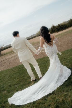 Couple in cream suit and lace wedding gown hold hands while walking on grassy hillside.