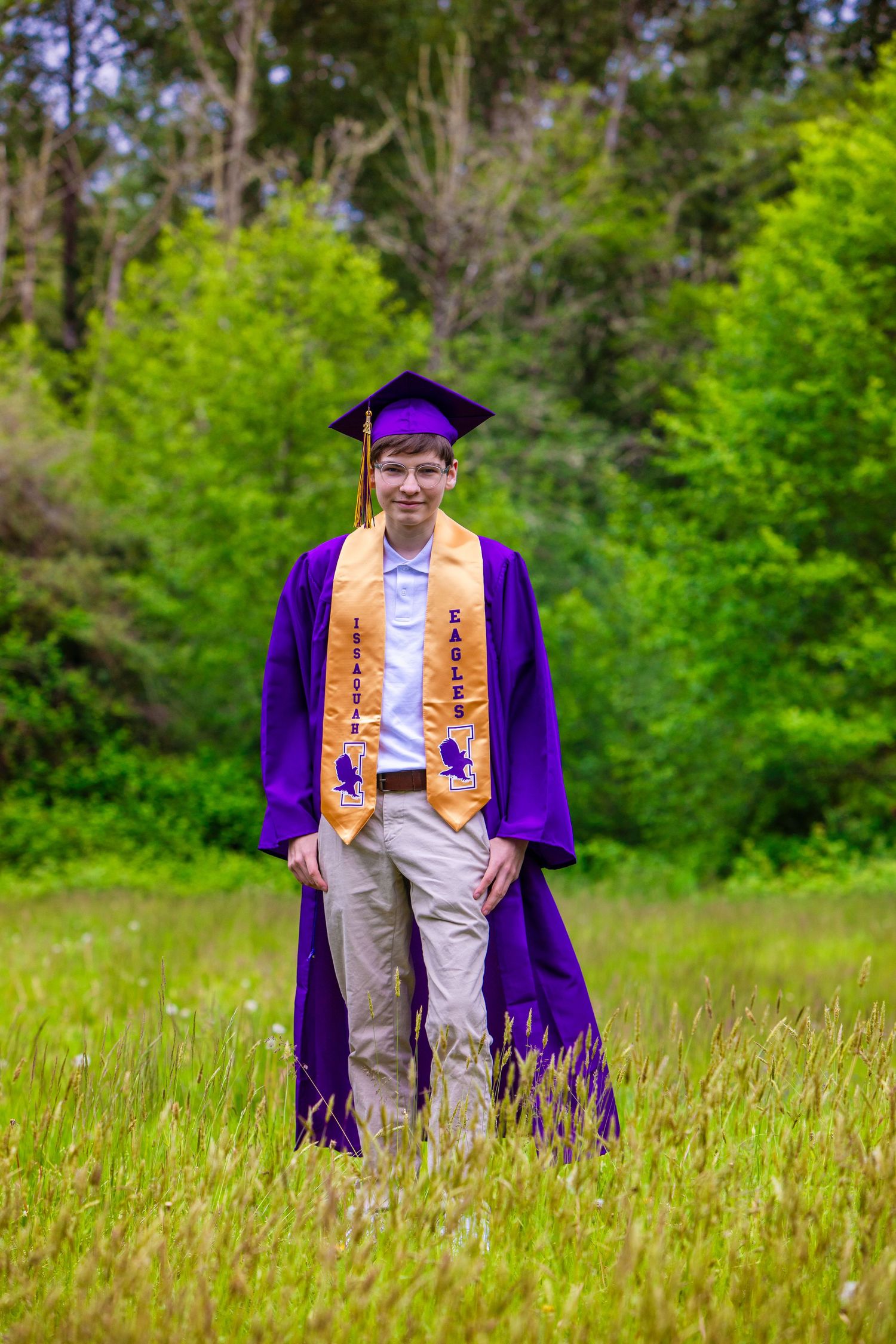 Graduate in purple and gold commencement regalia stands in a sunny meadow surrounded by green trees.