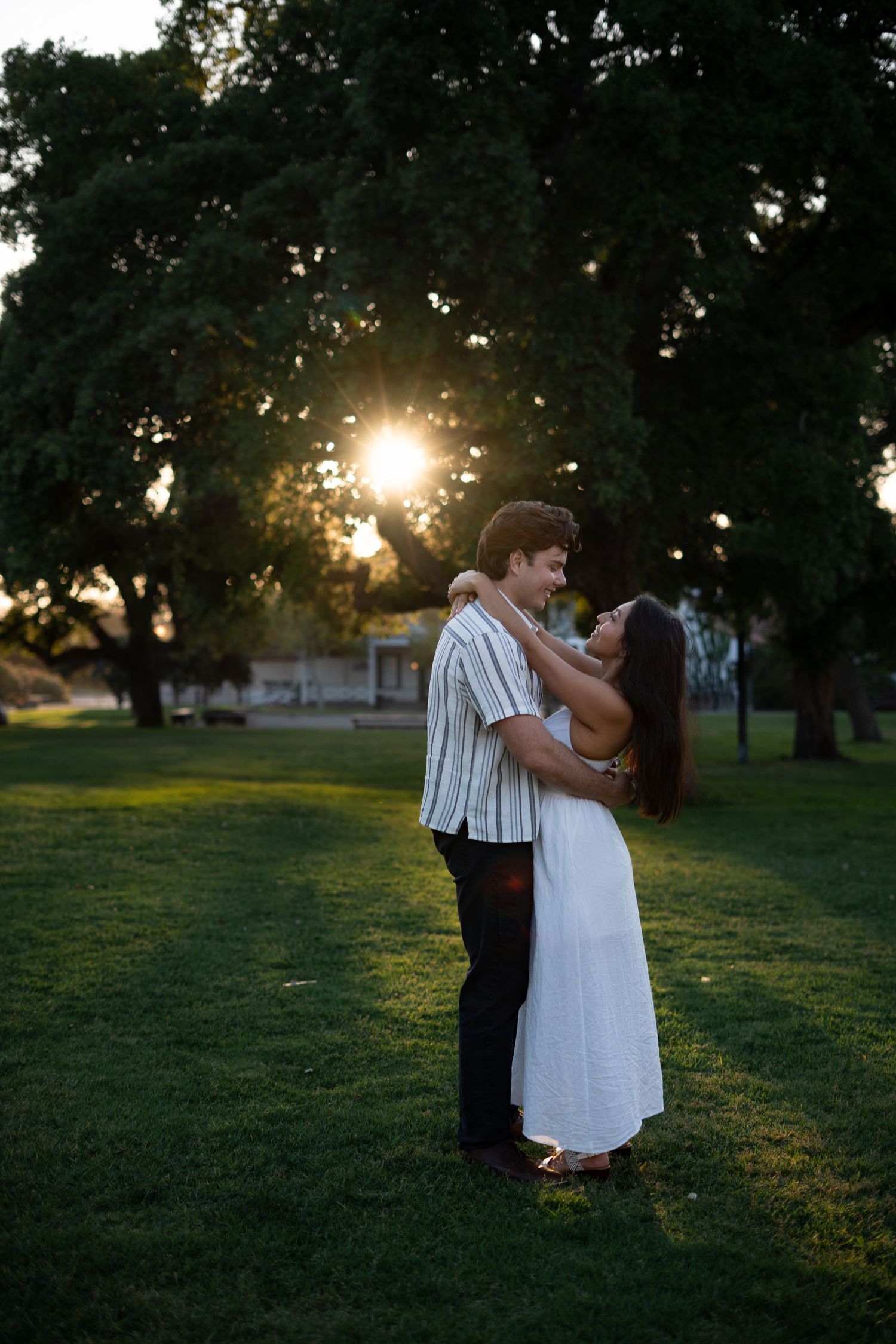 A couple shares a romantic moment at sunset in a grassy park with trees in the background.