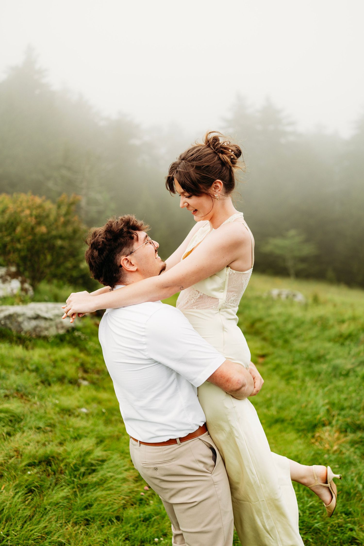 Man lifts smiling woman in yellow dress in a misty green mountain meadow surrounded by fog.
