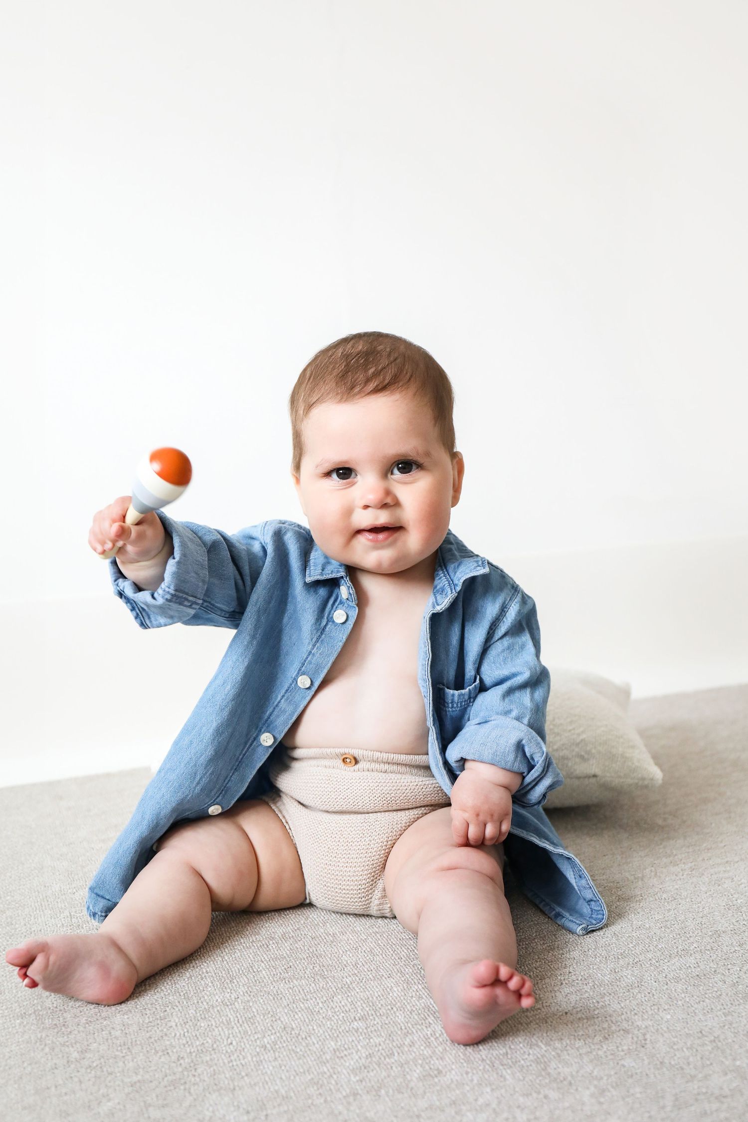Baby wearing denim shirt over beige shorts sits on grey surface while holding small orange toy, during Sitter photoshoot based in Nottingham