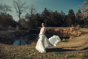A flowing white wedding dress billows dramatically in the breeze near a tranquil pond on an autumn afternoon.