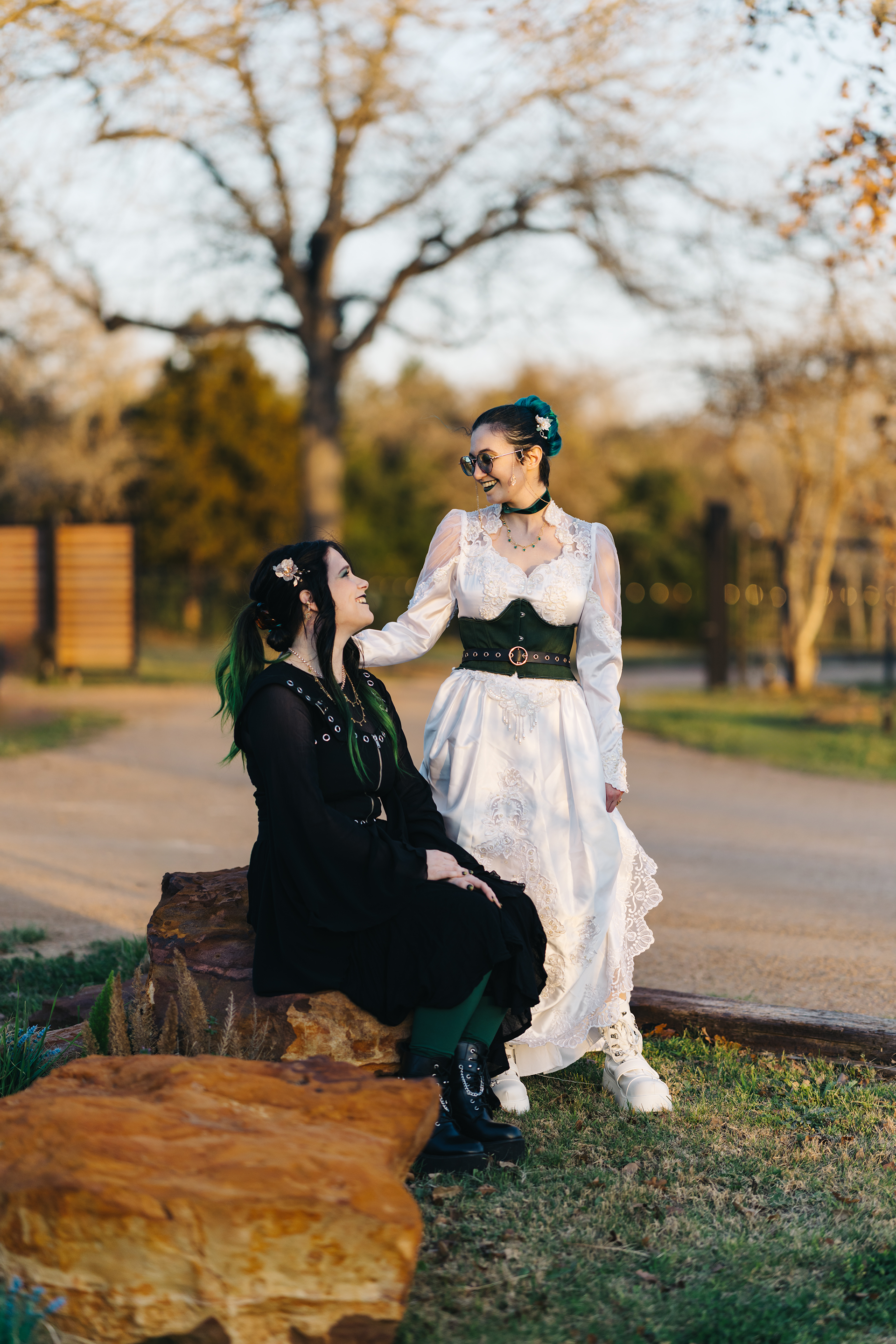 A couple sits on a rocky outcrop in a park during sunset, with one wearing a flowing white dress and the other in black.