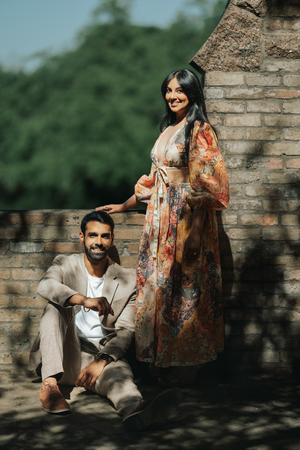 A couple poses near a brick wall in morning sunlight, with her wearing a floral dress and him in casual attire.