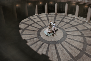 Aerial view of two people dancing in a circular courtyard surrounded by architectural columns casting dramatic shadows.