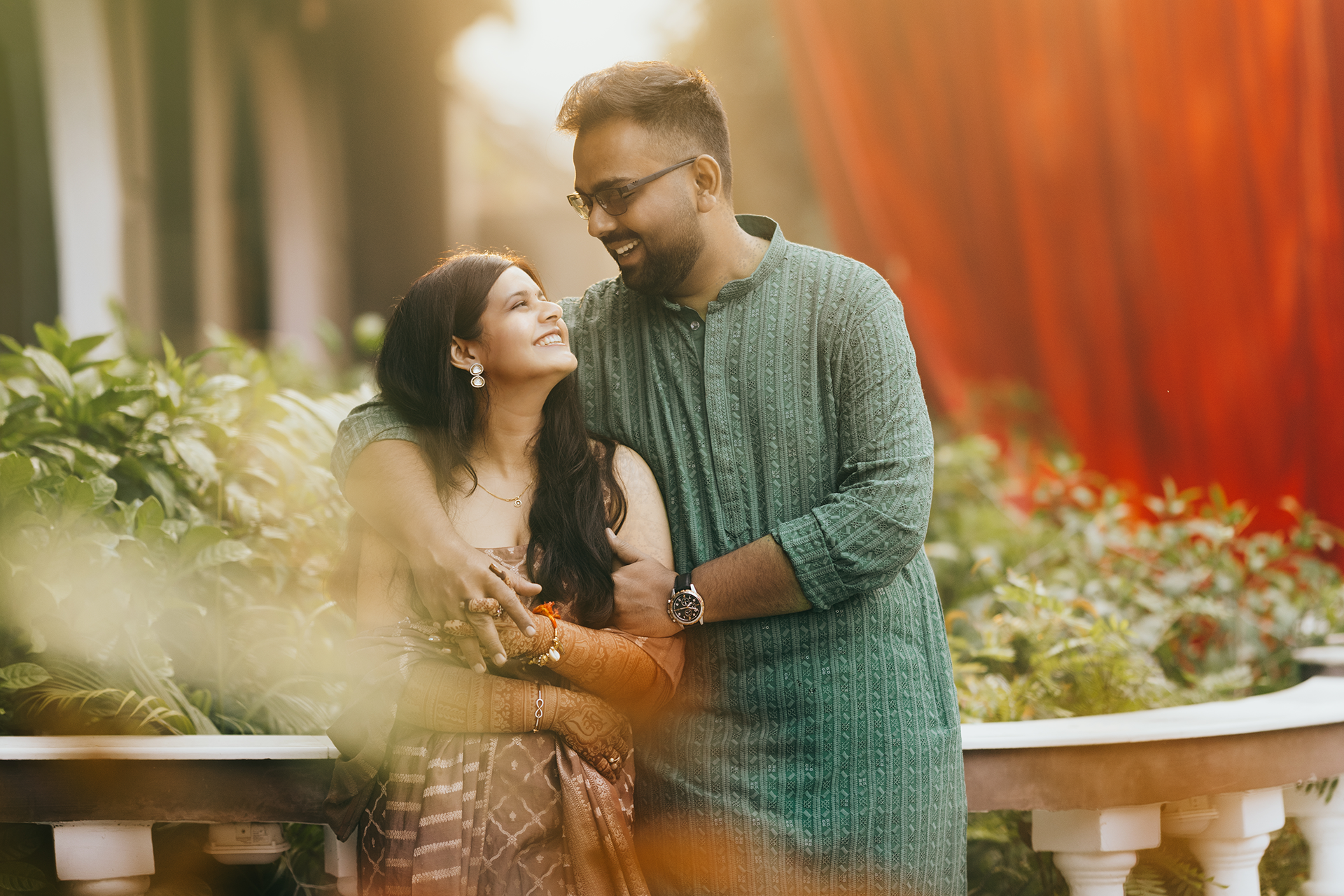 A couple shares romantic moments in a garden setting with warm autumn lighting and red foliage in the background.