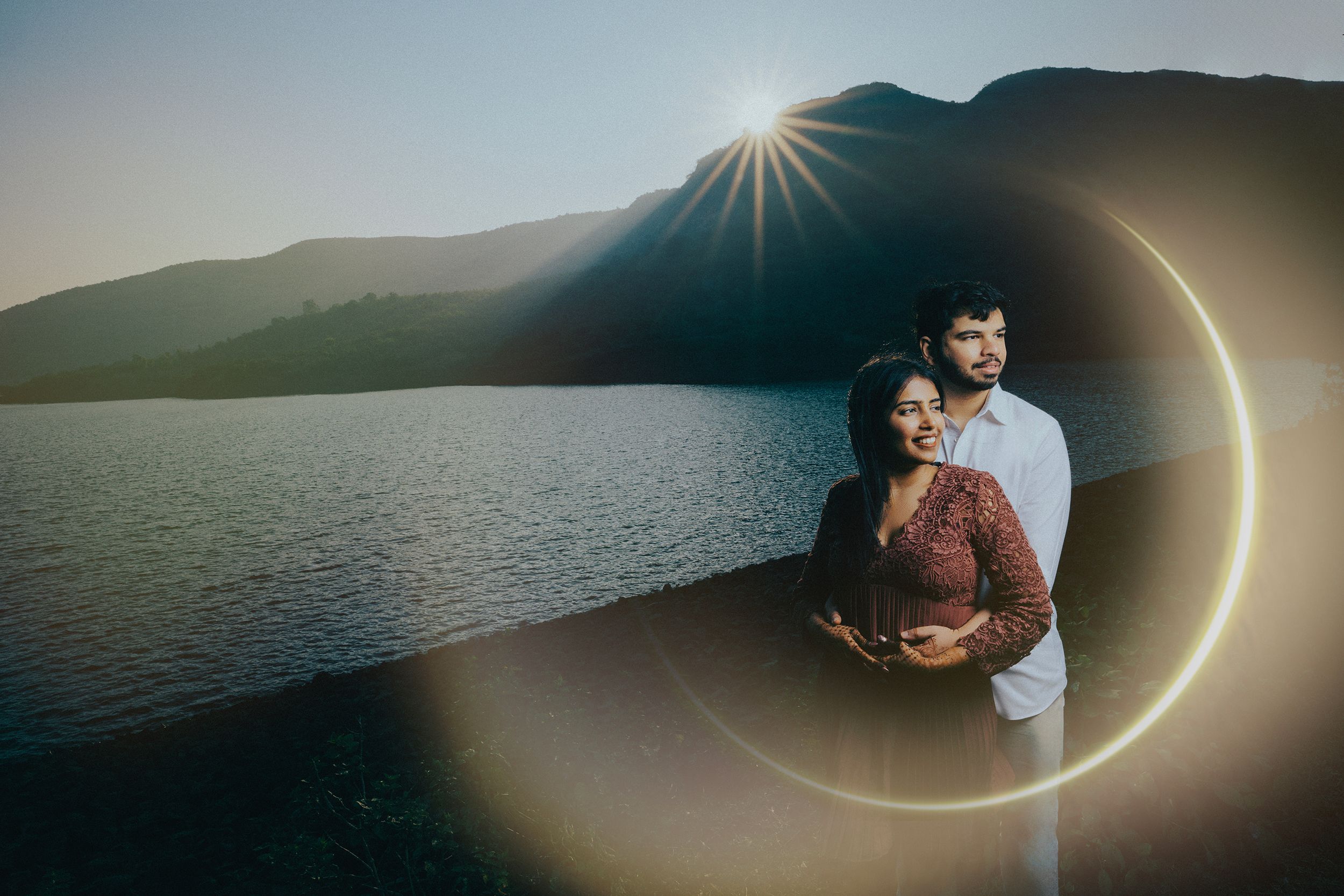 A couple embraces by a serene lake at sunset with sunburst and lens flare effects in the mountains.