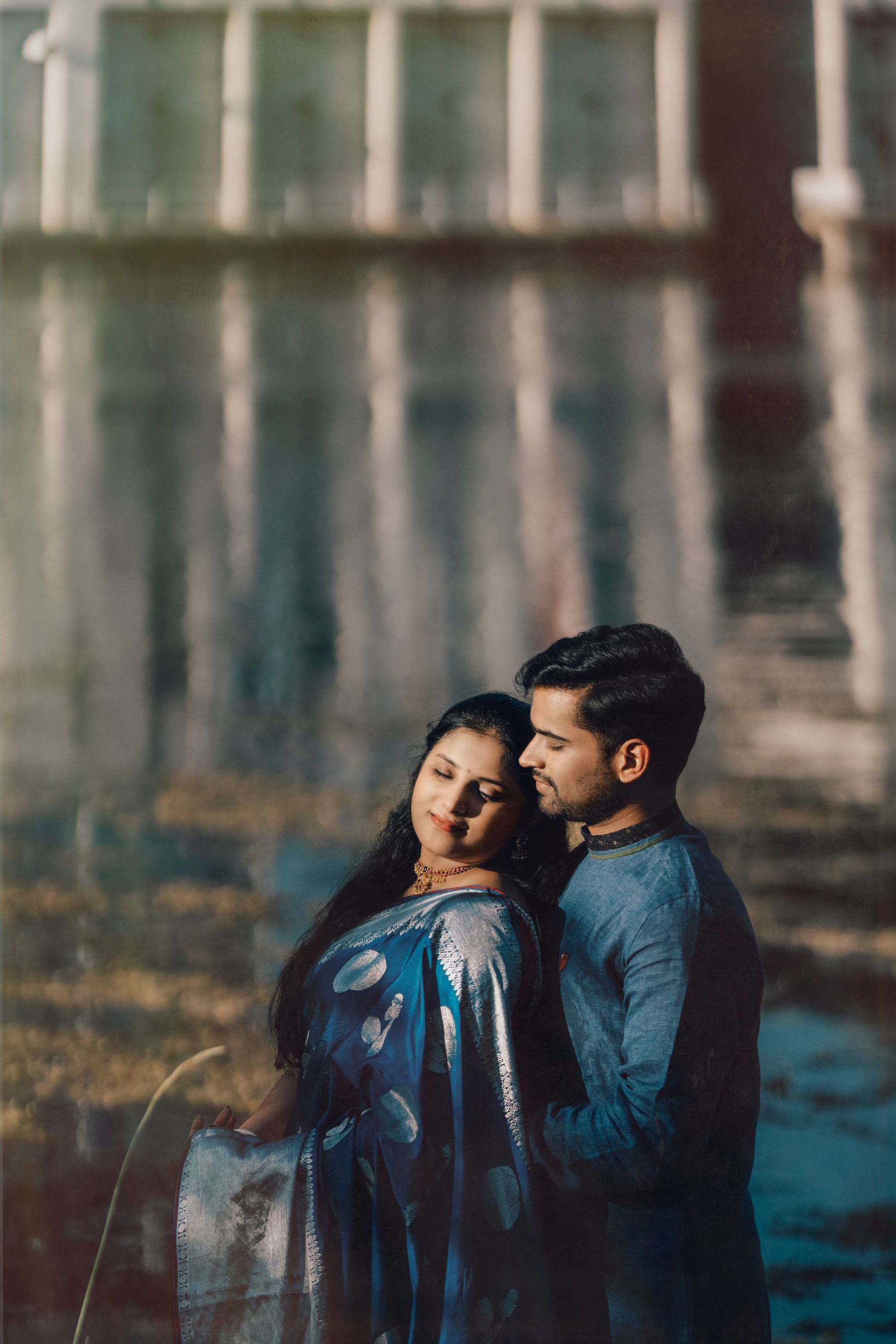 A romantic couple embraces outdoors, with the person wearing a blue patterned dress leaning against their partner in denim.