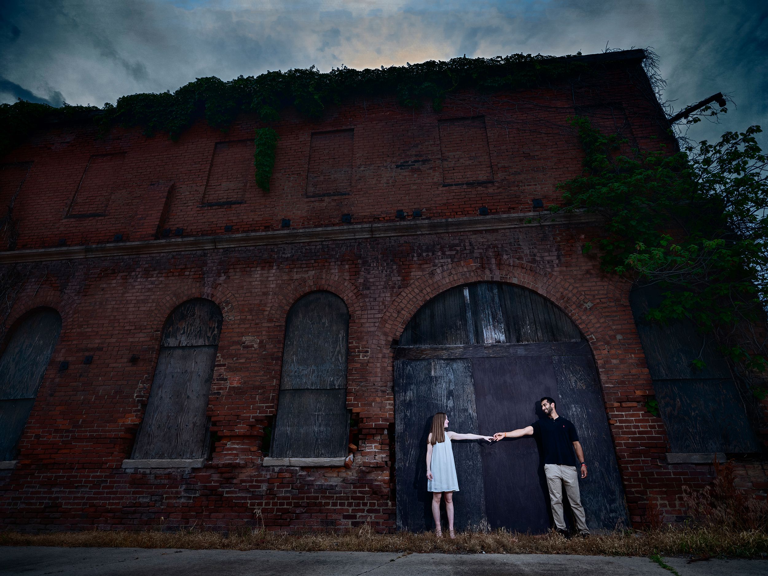 Two people stand in front of a moody old brick building with arched windows at dusk.