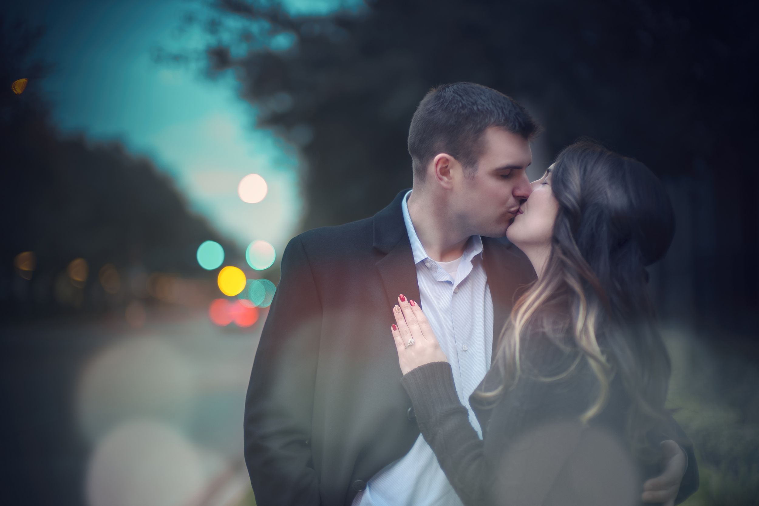 A romantic couple shares an intimate kiss at night with bokeh city lights creating a dreamy atmosphere in the background.