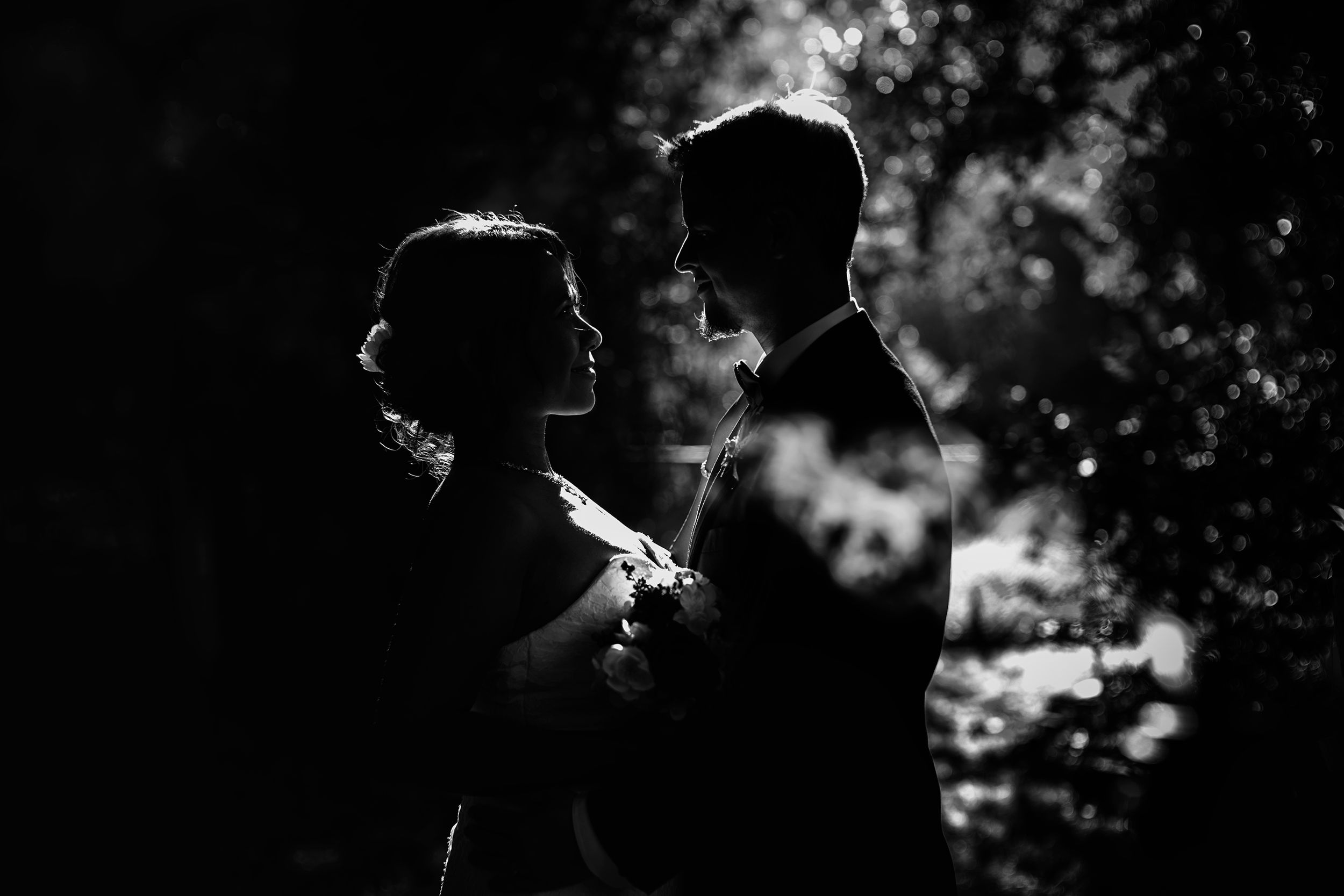 A romantic black and white silhouette of a couple standing close together in dappled sunlight through trees.