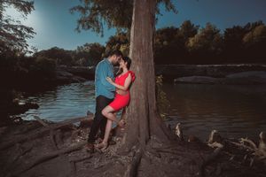 Couple shoot in blue and red embracing by a large tree trunk at sunset near a serene river.