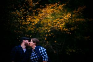 A couple stands close together and kiss in front of autumn foliage with golden leaves in soft evening light.