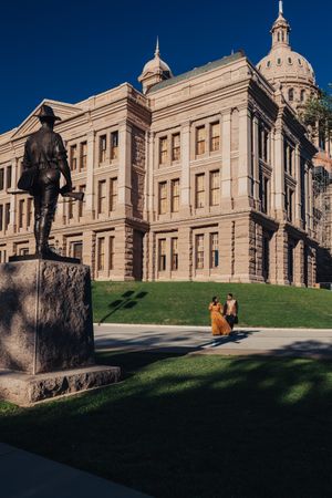 Historic Texas State Capitol building exterior with bronze statue and couple walking along landscaped grounds on sunny day.