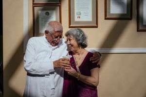 An elderly couple shares tender moments of laughter and affection together in a warmly lit room with framed photos on the wall.