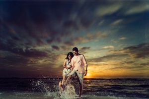 A couple shoot of the bride and groom having fun splashes in ocean waves during a dramatic beach sunset with clouds overhead.