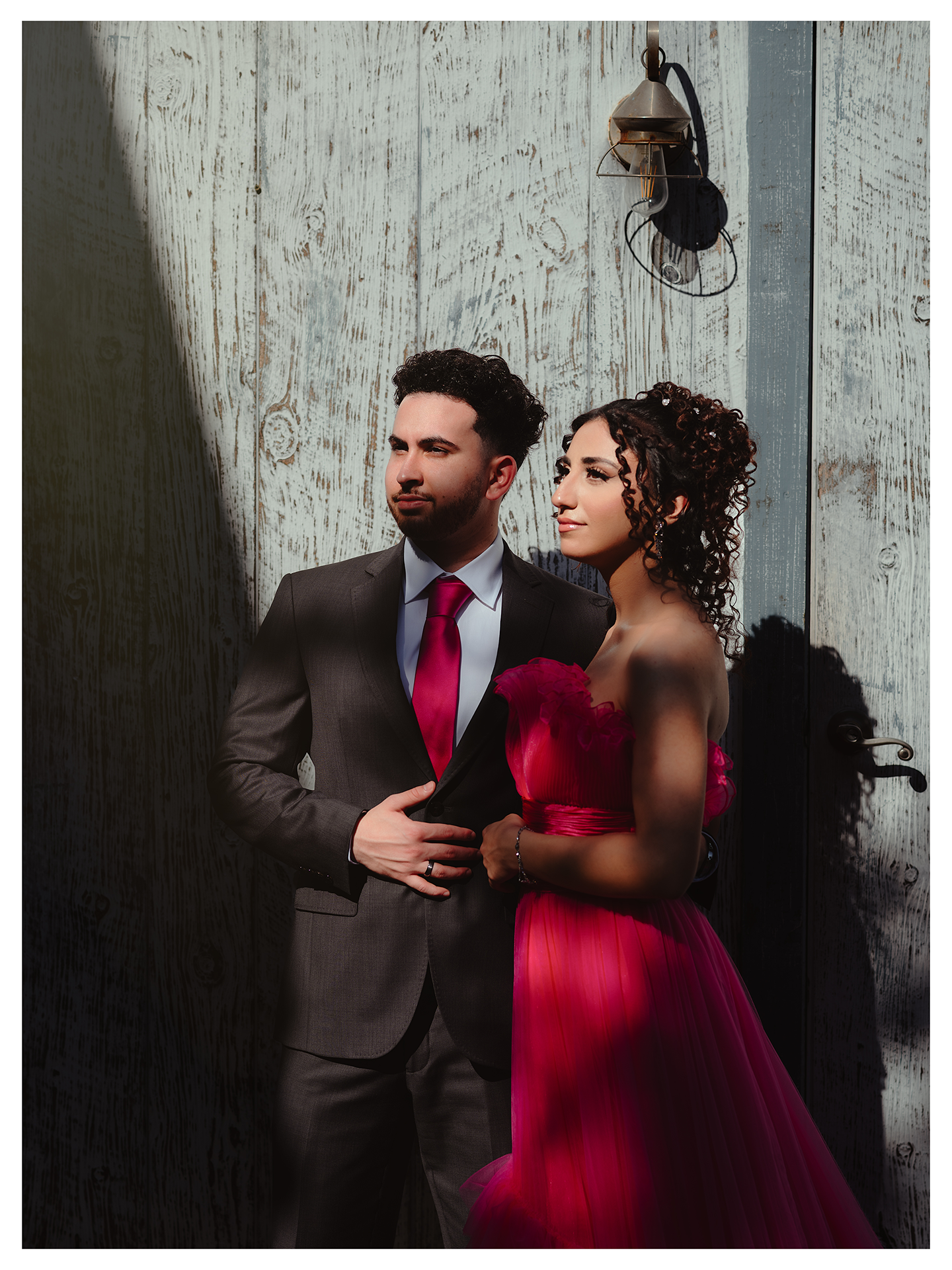 A couple poses against a rustic wall in formalwear - she in a stunning red gown, he in a grey suit with red tie.
