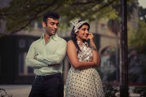 A couple poses outdoors in 70s Bollywood attire, with the woman wearing a polka dot dress and the man in a light button-down shirt.