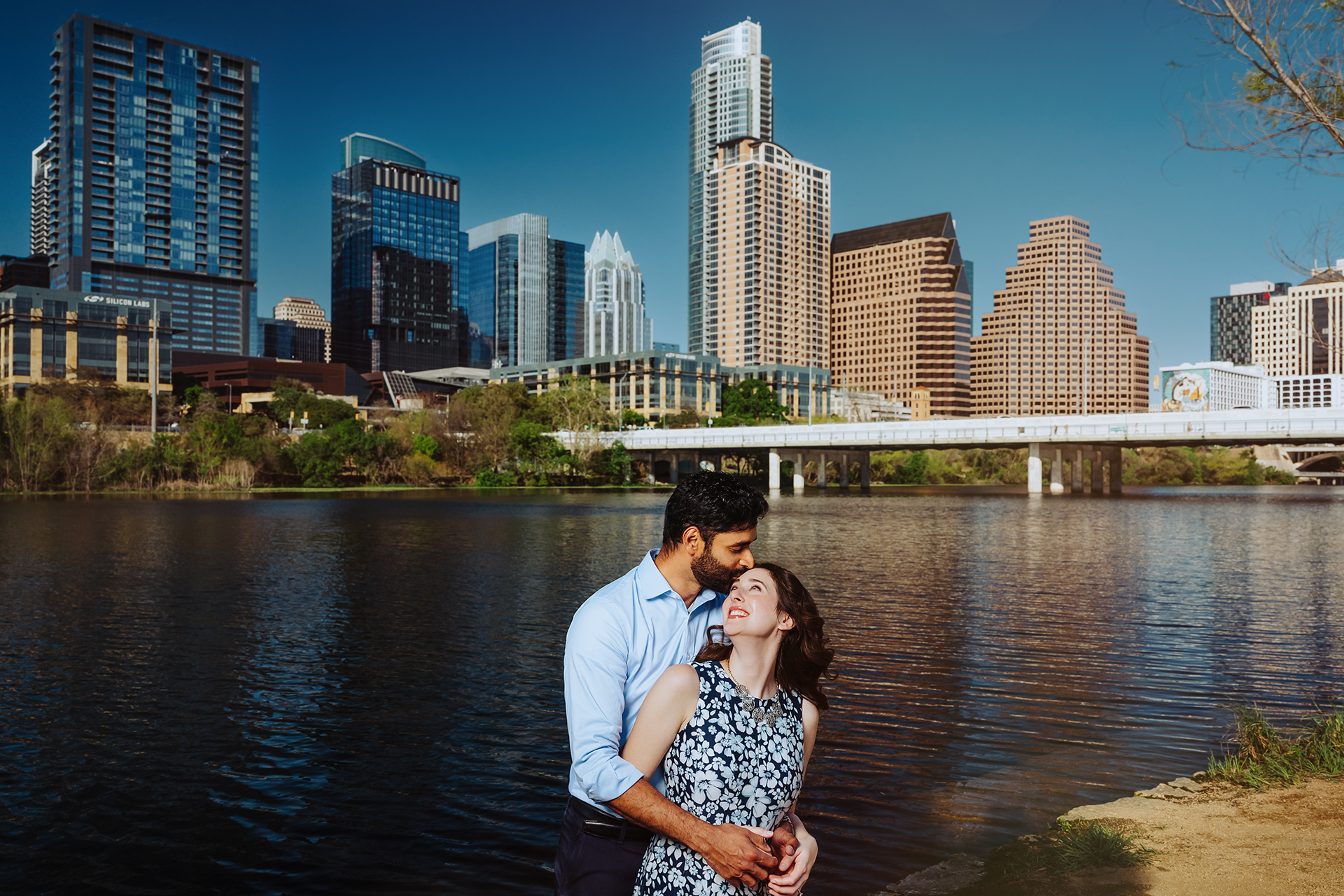 Couple embracing near Lady Bird Lake with Austin Texas skyline and bridge in background on sunny day.