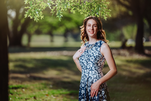 Young woman in floral blue dress posing outdoors in a sunlit park with lush green trees.