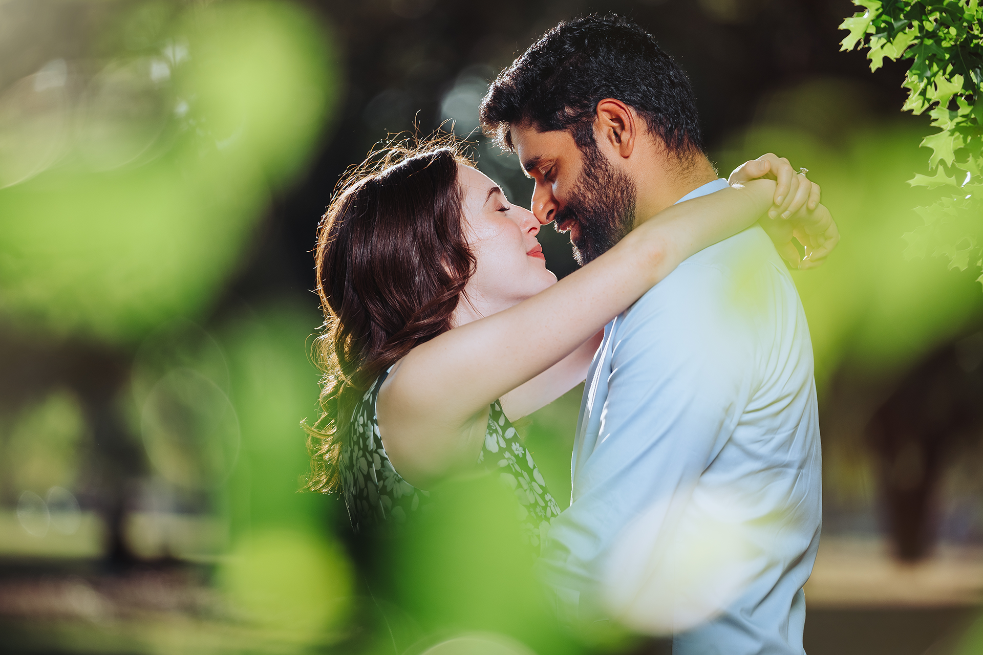 Romantic couple embracing and almost kissing outdoors in a lush green park setting on a sunny day.
