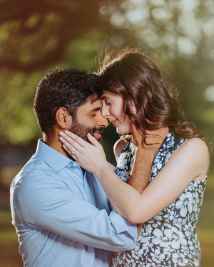A romantic couple shares an intimate moment outdoors, foreheads touching, surrounded by soft bokeh greenery.