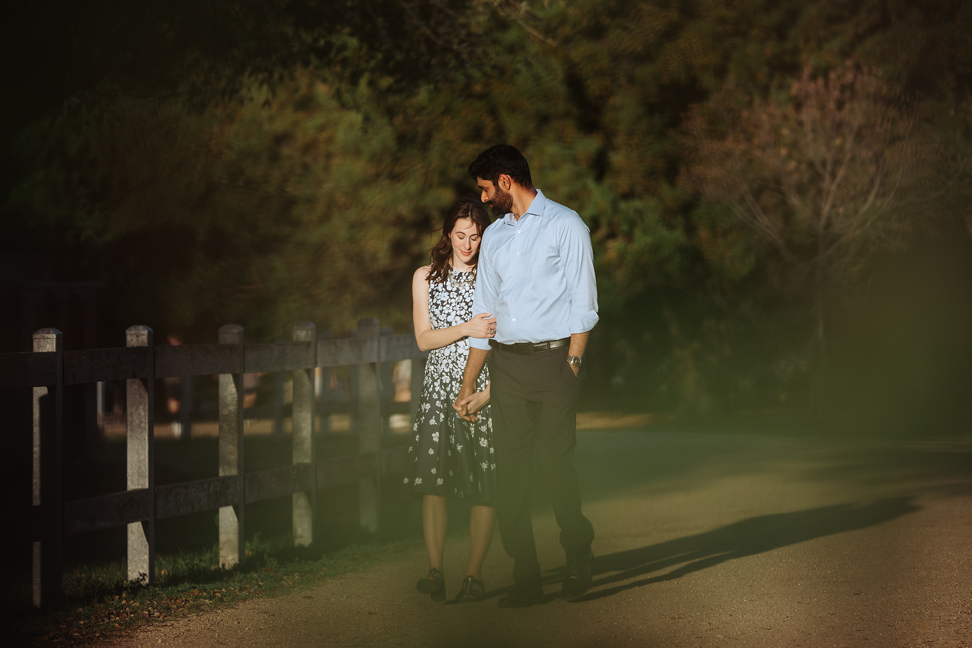 Couple standing close together on a quiet road at night surrounded by lush green trees in a romantic outdoor setting.