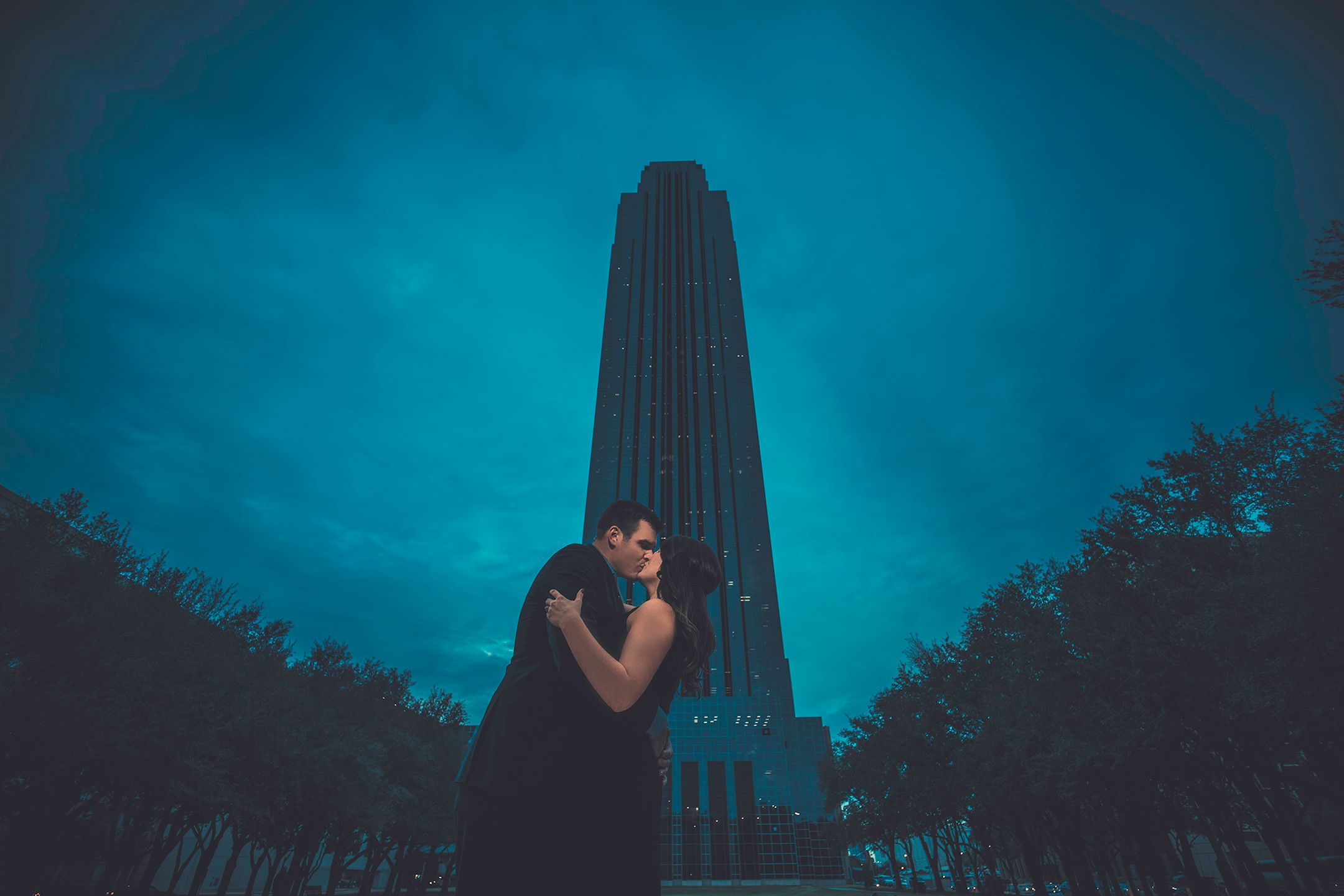 A romantic silhouette stands against a tall skyscraper tower under a moody teal night sky.