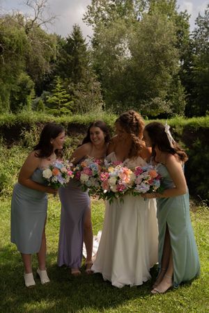 Bridal party in pastel dresses huddle together while holding colorful bouquets in a garden setting.