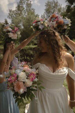 A bride in a white dress poses with colorful floral bouquet and crown against a cloudy sky.
